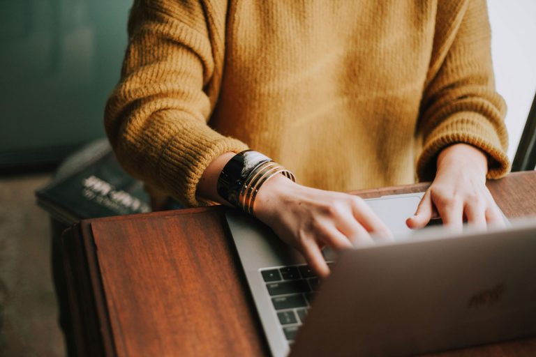 Eine Person mit goldfarbenem Pullover und auffälligem Armreif tippt konzentriert auf einem Laptop. Der Fokus liegt auf den Händen und dem Gerät – nicht auf dem Gesicht. Die Szene spielt sich an einem Holztisch ab, im Hintergrund liegt ein schwarzes Notizbuch mit der Aufschrift „Write What You Know“. Das Bild symbolisiert kreatives Arbeiten, digitale Textproduktion und den bewussten Umgang mit Sprache – passend zum Thema inklusives und barrierefreies Schreiben.