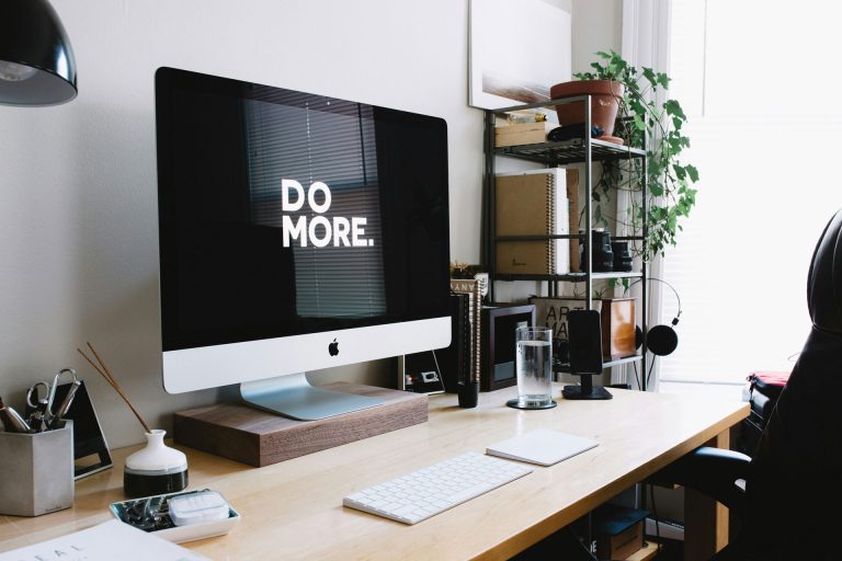 Minimalistischer Arbeitsplatz mit einem großen Apple-Computer, auf dessen schwarzem Bildschirm in weißer Schrift „DO MORE.“ steht. Der Schreibtisch aus hellem Holz ist ordentlich eingerichtet mit Tastatur, Maus, Wasserglas, Headset und Schreibutensilien. Im Hintergrund stehen Notizbücher, Pflanzen und Bücher im Regal. Das Bild vermittelt eine aufgeräumte, kreative Arbeitsatmosphäre und passt zum Thema strategischer Content und wirkungsvolle Blogkommunikation.