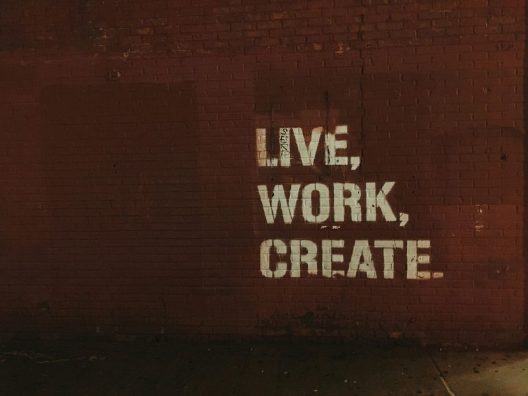 Rote Backsteinwand mit weißem Schriftzug in Versalien: „LIVE, WORK, CREATE“. Die Worte sind im Stil von Streetart oder Graffiti aufgesprüht und leuchten vor dem dunklen Hintergrund. Die Aufnahme wirkt urban, kreativ und motivierend – passend zum Thema Zusammenarbeit, Kreativität und moderne Texterstellung.
